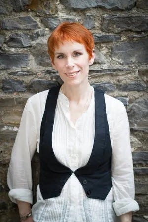 author photo of tana french; shock red pixie cut, white blouse, effortlessly cool black vest thing over it, gah she's amazing