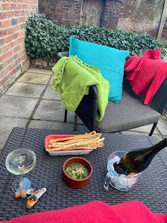 A garden table with chairs either side of it. On the table is a bottle of wine in a makeshift icebucket (a pyrex jug with ice cubes) a platter of bread sticks, a bowl of green dip (pesto) and a glass of wine.