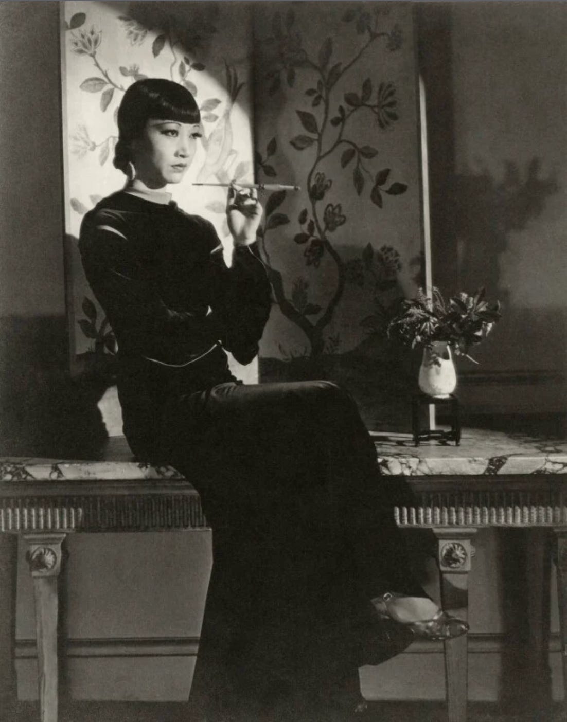 Anna May Wong sits on a marble mantel in a long, black velvet dress with a cigarette in a long holder held to her face