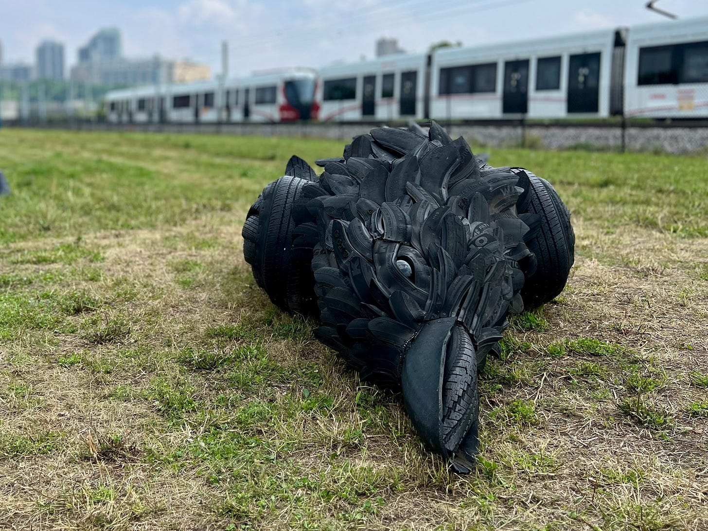 A five metre long dead crow, made of used car tires, lying on the grass with the Ottawa LRT train in the background.
