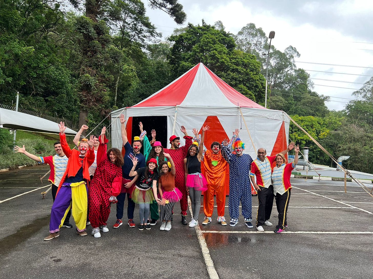 Foto de diversos jovens com roupas de palhaço, bailarina e outras fantasias em frente a uma tenda de circo.