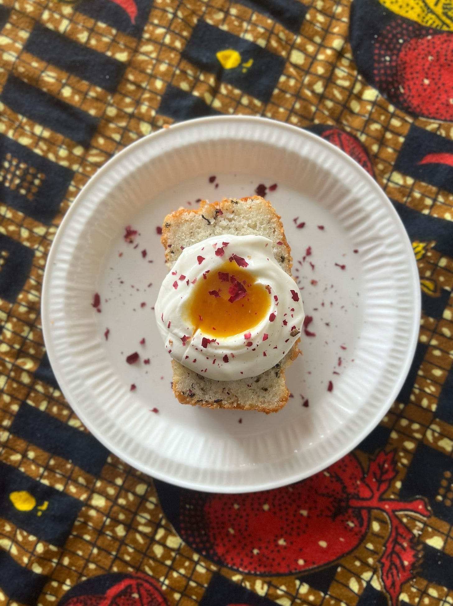 multicolor ankara tablecloth underneath a white plate with a slice of cake topped with a dollop of yogurt and passion fruit with dried rose petals 