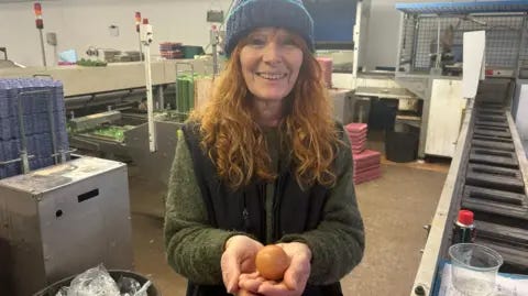 BBC Alison Greene pictured in the processing unit with a conveyer belt running next to her. She is wearing a green jumper with a black woolly gilet and a blue woolly hat. She has red hair and is holding the brown egg in front of her.