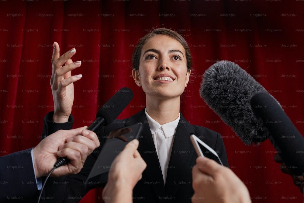 Waist up portrait of smiling female official giving interview to journalist and speaking to microphones against red curtain