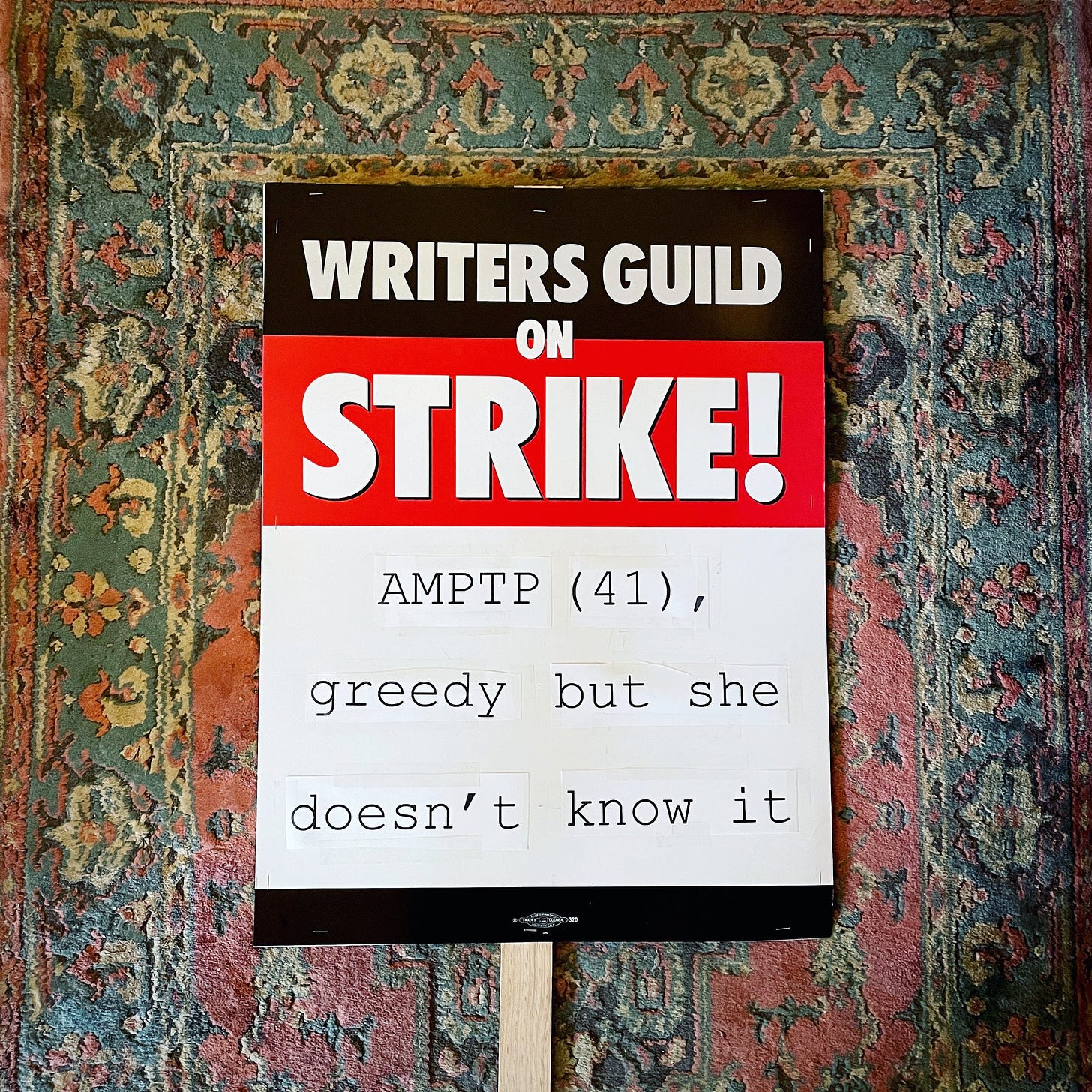 a "writers guild on strike" picket sign on a rug; the bottom half says "AMPTP (41), greedy but she doesn't know it"