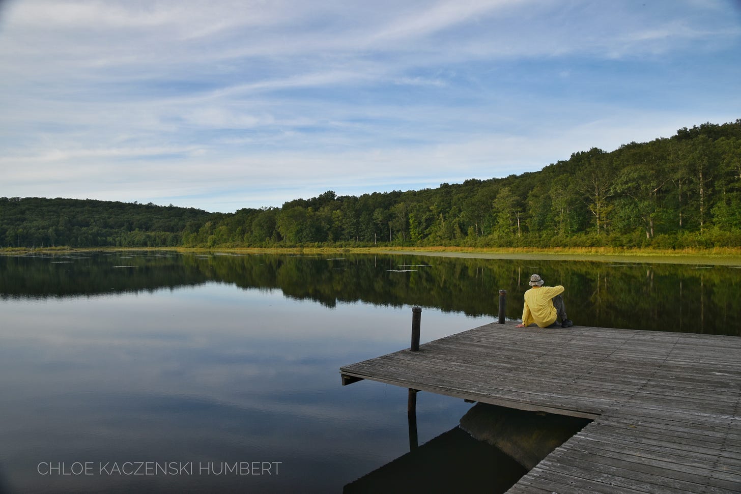 Cobey Pond, Pennsylvania State Gamelands 316, August 26, 2019. Photo by Chloe Kaczenski Humbert. Someone sitting on a dock at a quiet pond where the water is still and the shoreline is wild and there is no sign of people or structures.