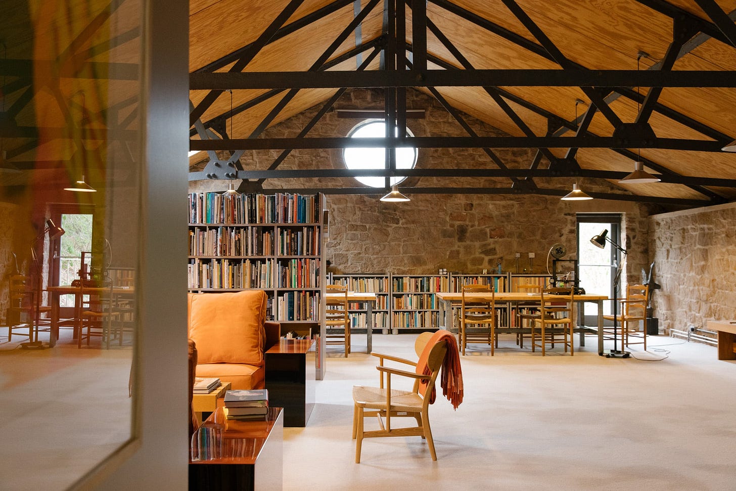 A living room with a vaulted ceiling and exposed steel frame. Bookshelves to the back of the room, mid-century oak tables and chairs. 