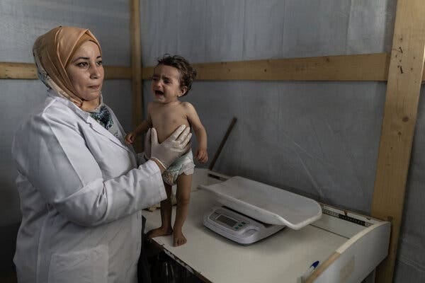 A person in a white coat tends to a small, thin child standing on a tablelike surface near a scale. A person in a white coat tends to a small, thin child standing on a tablelike surface near a scale.
