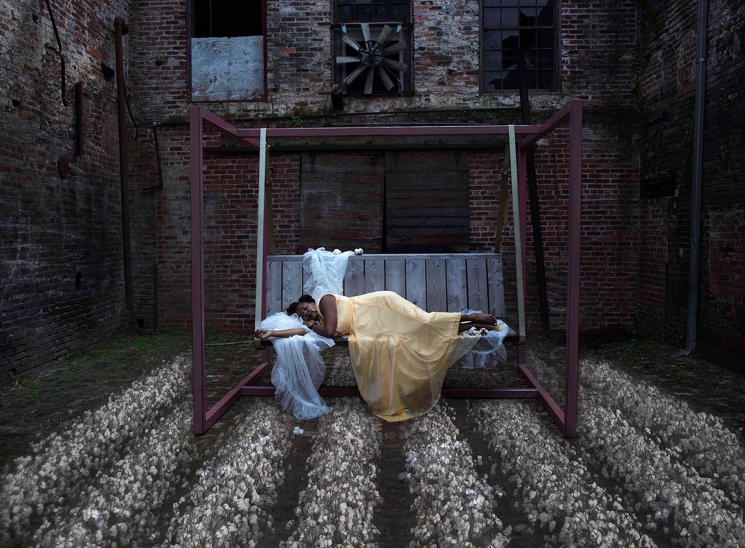 Photo of Tricia Hersey in a yellow gown napping on a bench in an urban landscape over rows of cotton; Credit: Charlie Watts.