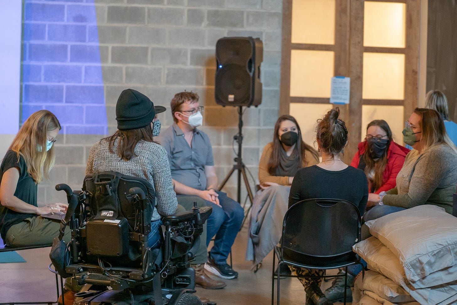 A group of mostly or entirely white people, including a powerchair user, are seated in a circle of chairs wearing KN95 masks. A pile of pillows is nearby.