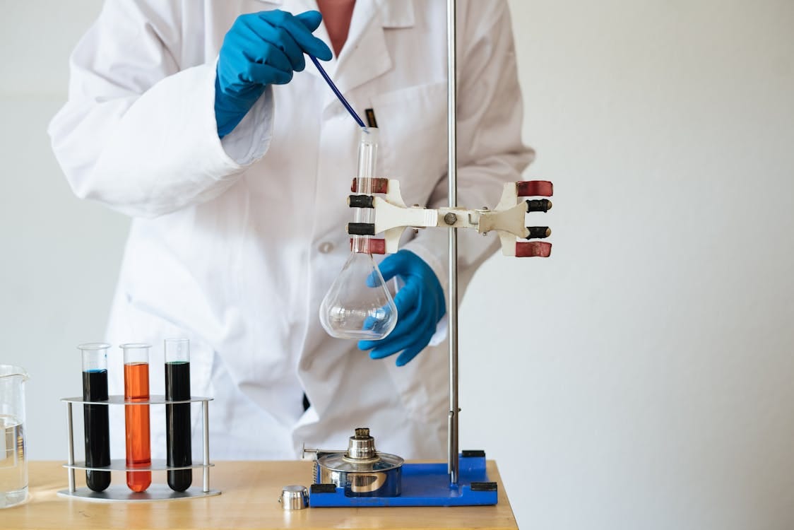 Free Crop of unrecognizable scientist wearing lab coat and gloves and inserting pipette into empty flask mounted on ring stand while working in laboratory Stock Photo