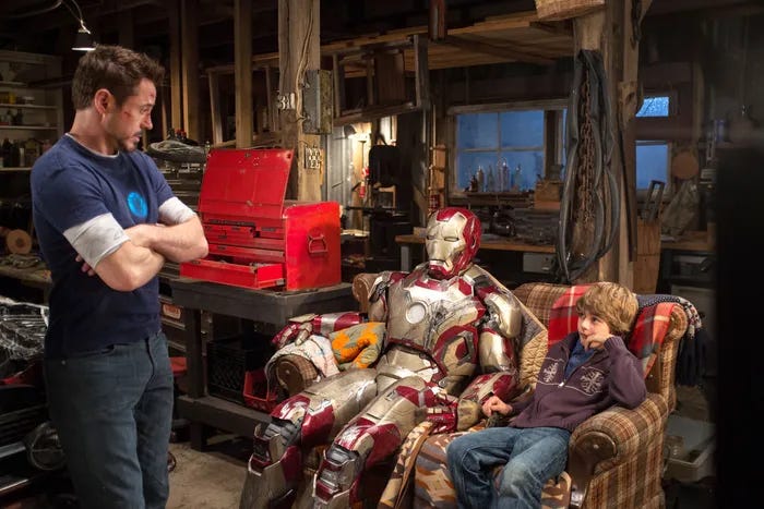 A screenshot from Iron Man 3 showing Tony Stark looking at his Iron Man suit and a child sitting which are both sitting on a couch.