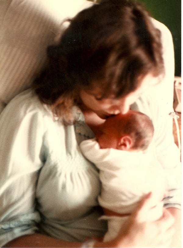 A mother in a nightgown holds a tiny baby in her arms and kisses his head.