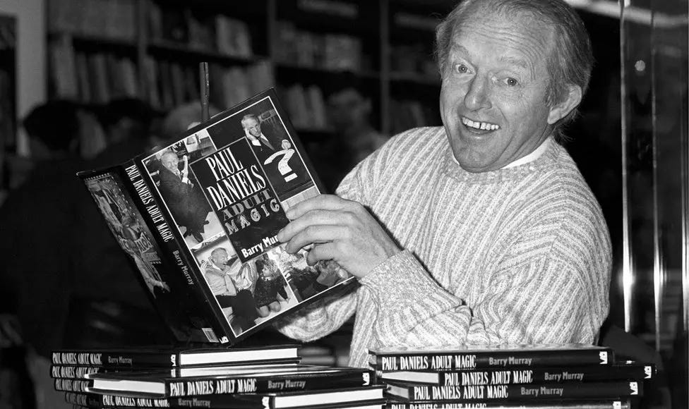 Black and white photograph of magician Paul Daniels, an older white man, holding up a magic book at a book signing event Black and white photograph of magician Paul Daniels, an older white man, holding up a magic book at a book signing event