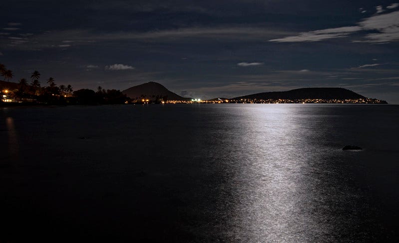 Muted night-time colors show moonlight reflecting off Maunalua Bay on the shoutheast shore of Oahu, with houselights showing at the base of Koko Head crater in the background, and palm tree silhouettes on the right-hand side of the photo in the background.