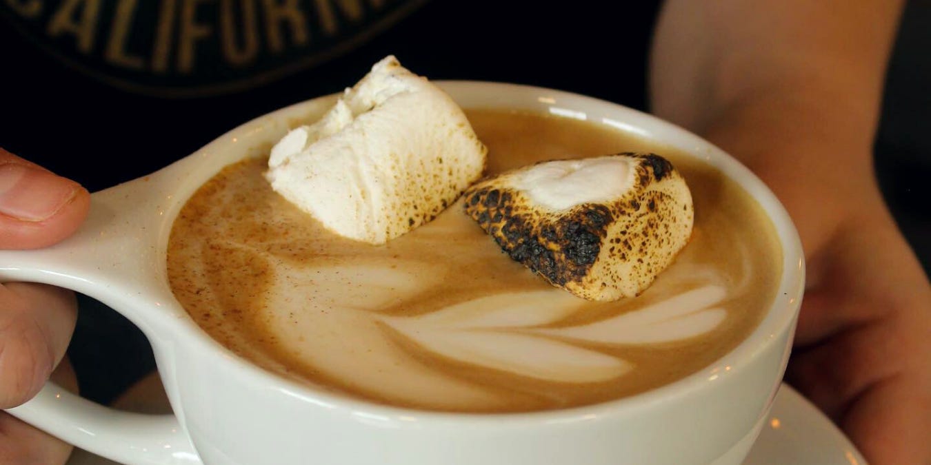 A close-up of flower latte art with two toasted marshmallows floating in a latte coffee. The mug is traditional porcelain white.