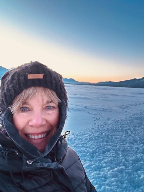 Woman bundled up in winter clothes, a snow covered lake and mountains in the background with the sun rising.