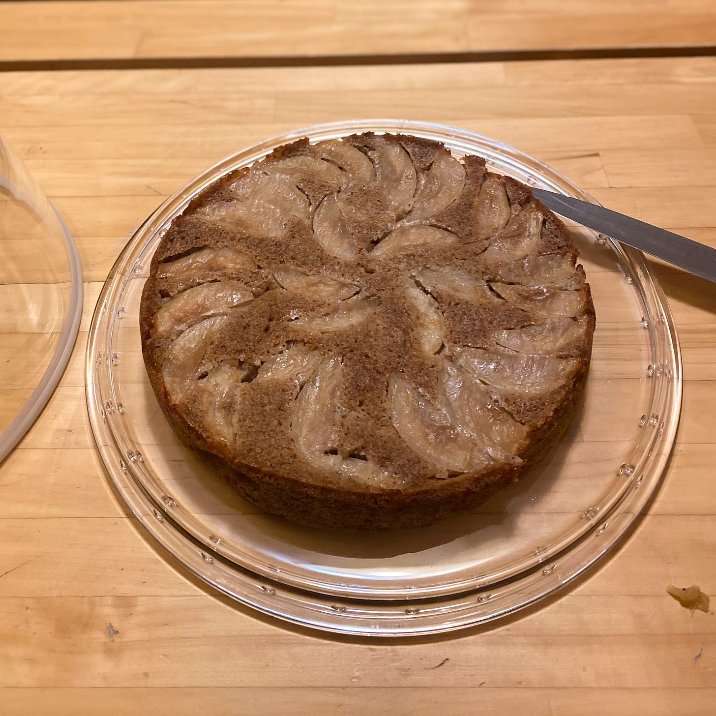 An apple upside down cake on a clear glass cake plate, slices of apple arranged over the top. A knife rests on the side of the plate.