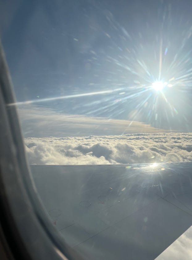A photo taken out of the window of a plane. You can see the wing and the edge of the window. Beyond this is a bright starburst of a sun, blue sky, and a floor of textured, fluffy clouds.