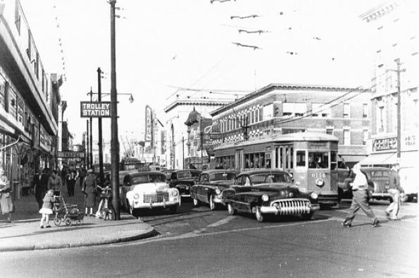 Black and white photo of a crowded intersection with a trolley and cars from the 1950s.