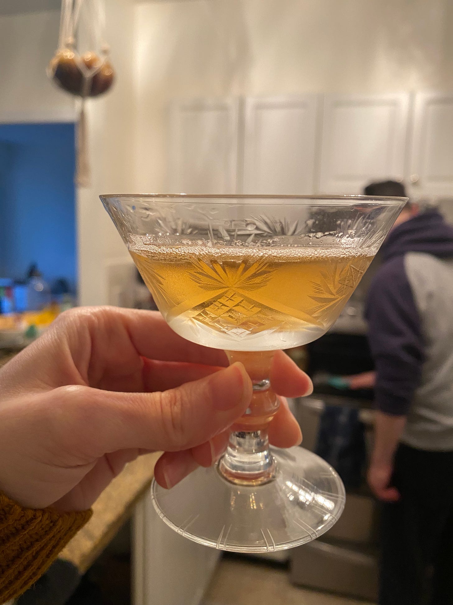 The etched glass described above, with a stylized pineapple visible in the design. I'm holding it up in the kitchen, it's about 3/4 full with a honey-coloured drink. Jeff is wiping the stove in the background, wearing a blue and grey hoodie.