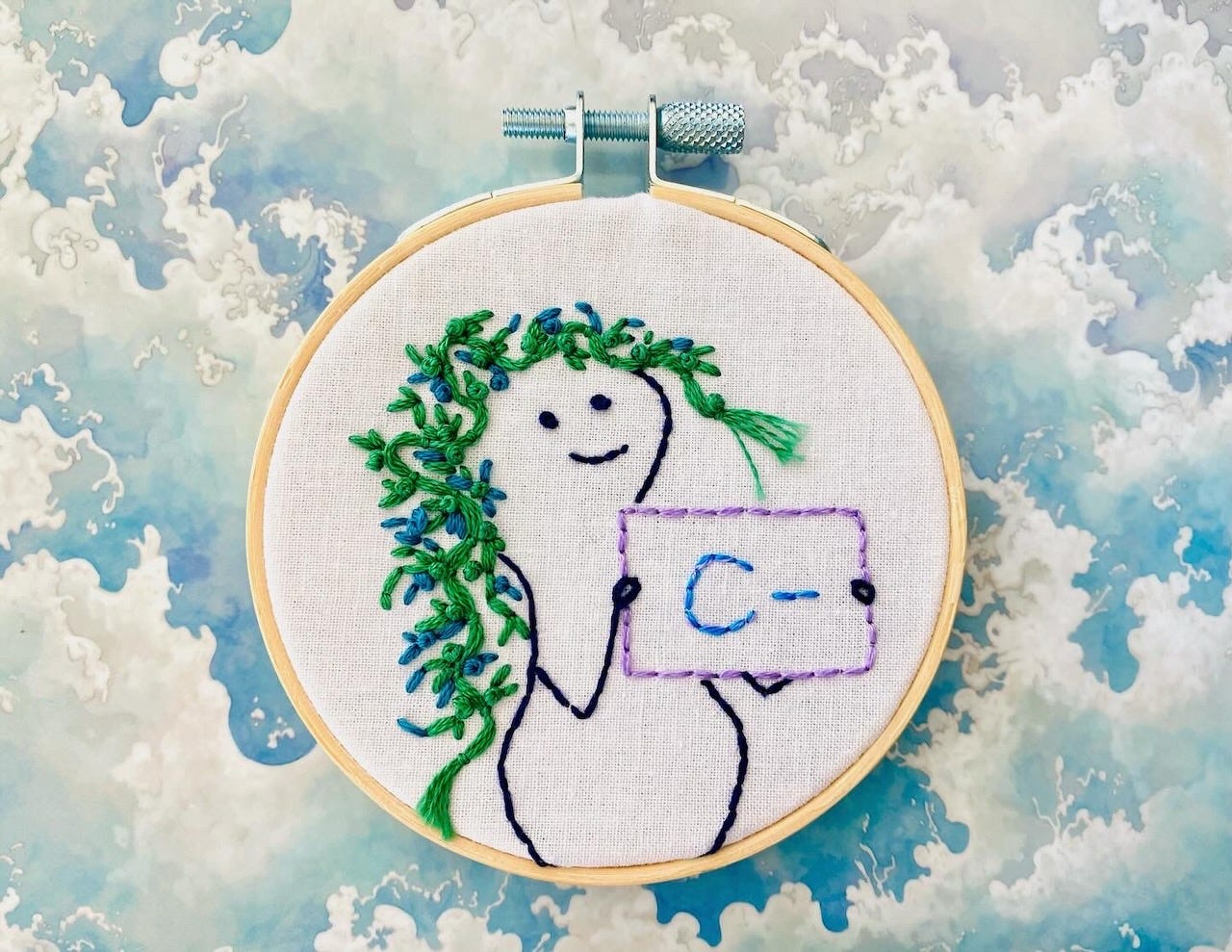 Miniature embroidery of a peanut-shaped figure with luxurious blue-green seaweed hair holding up a sign that says “C-.:”