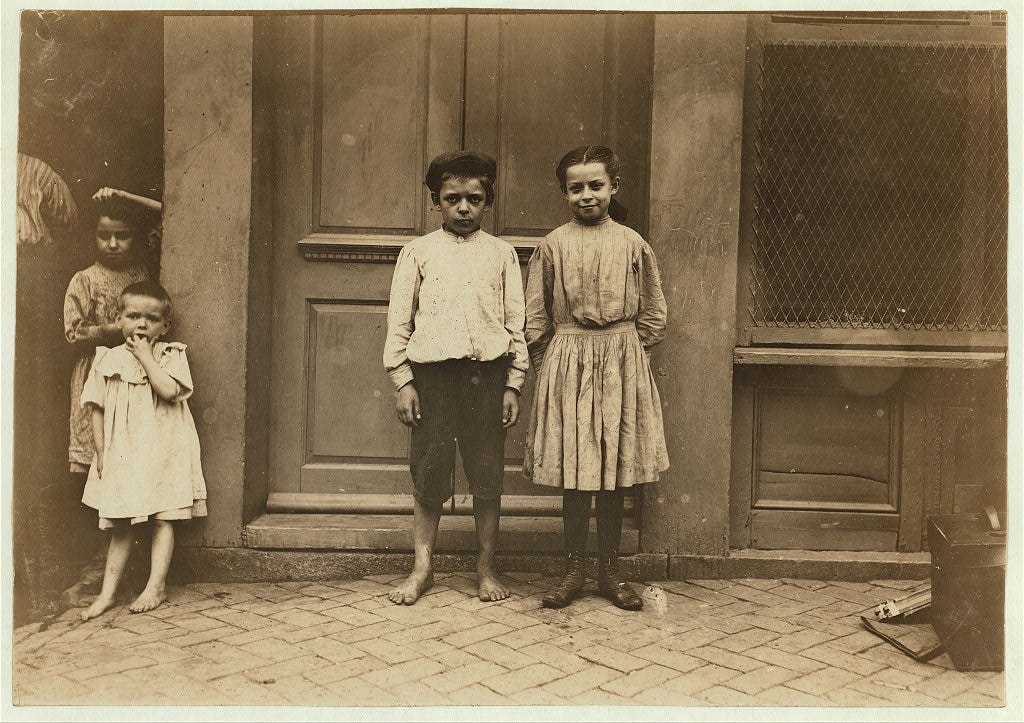 A sepia-toned photograph of two white children in working clothes taking a break from working jobs as child laborers. They look tired.