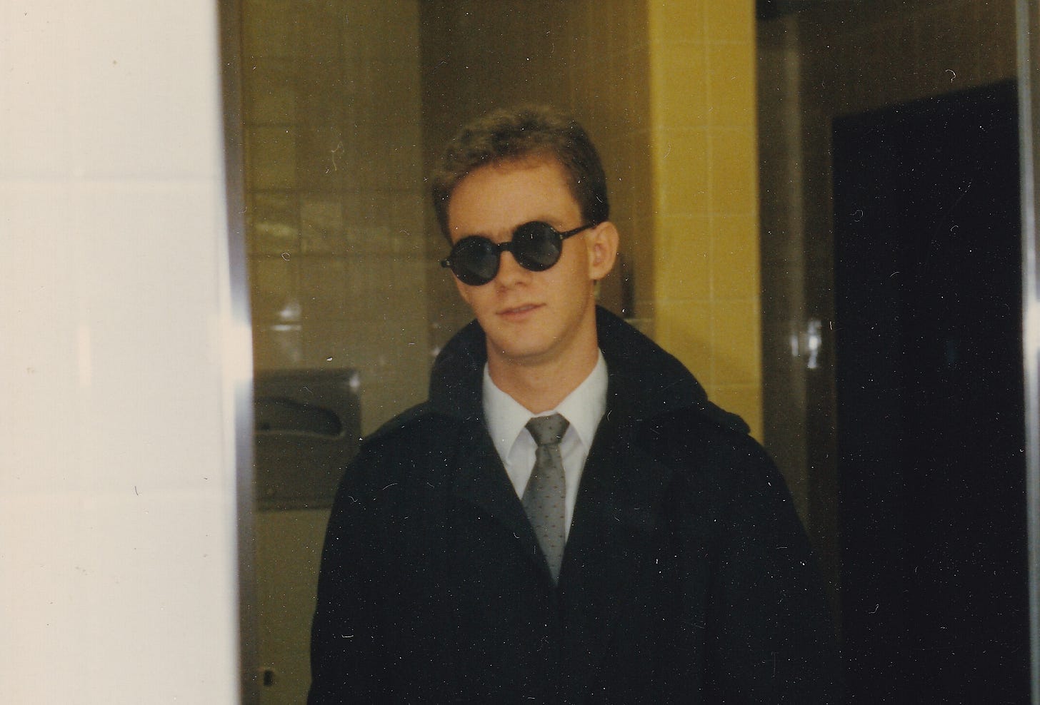 A sneering young man in a suit, black trenchcoat, and dark round sunglasses sneers into a mirror in a yellow-tiled restroom.