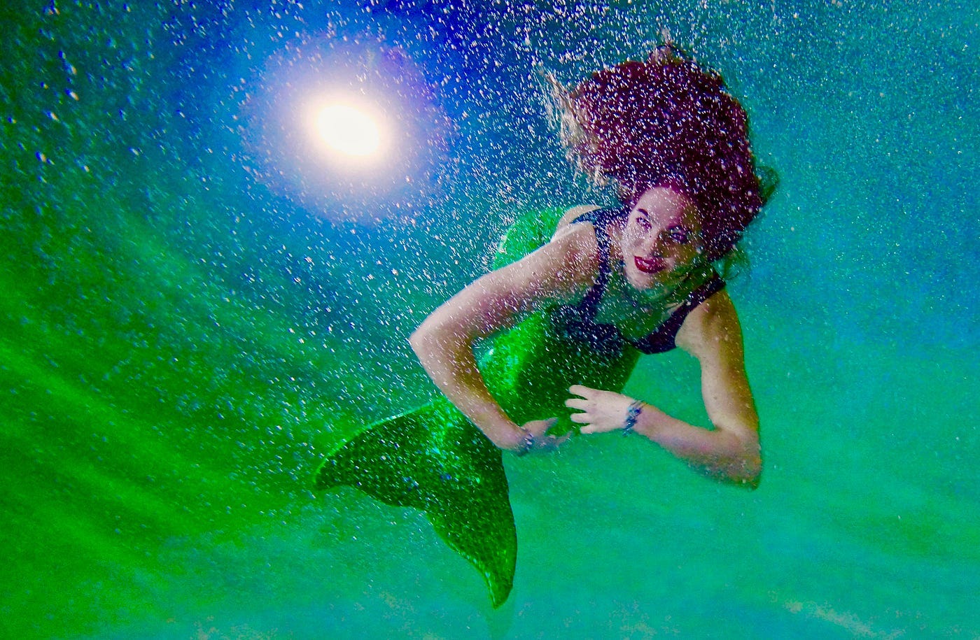 A woman wearing a mermaid fin and bathing suit is submerged in the sea bottom. The glowing round moon shines in the background surrounded by starts. The woman faces the viewer, her long and full red hair swaying towards the middle top of the frame. The woman looks happy.