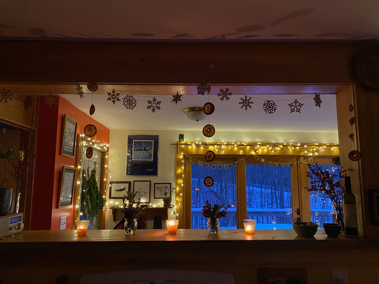 View through an opening. from my kitchen into my dining room. The countertop is decorated with lit candles and jars of winterberry; wooden snowflakes and dried orange slices hang from the ceiling beam; the windows are lined with twinkly lights.