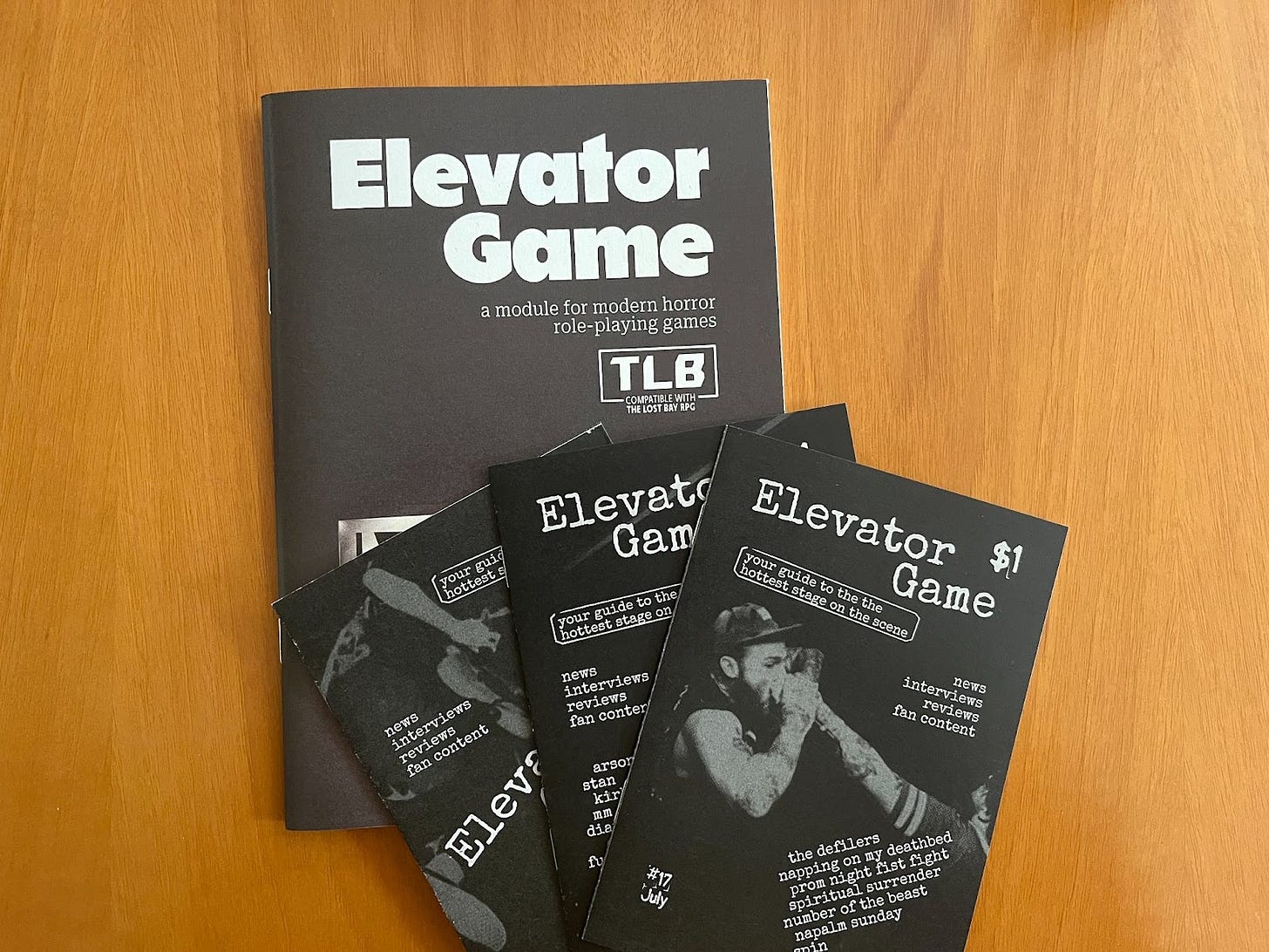 The zine Elevator Game, designed by Allen Hall, compatible with the Lost Bay, as well as three player-facing mini-zines. The covers are black and white. The main zine features an elevator display, showing that the first floor is below and the second above. The three mini-zines are styled after homemade punk zines, and claim to contain “News, interviews, reviews, and fan content.”