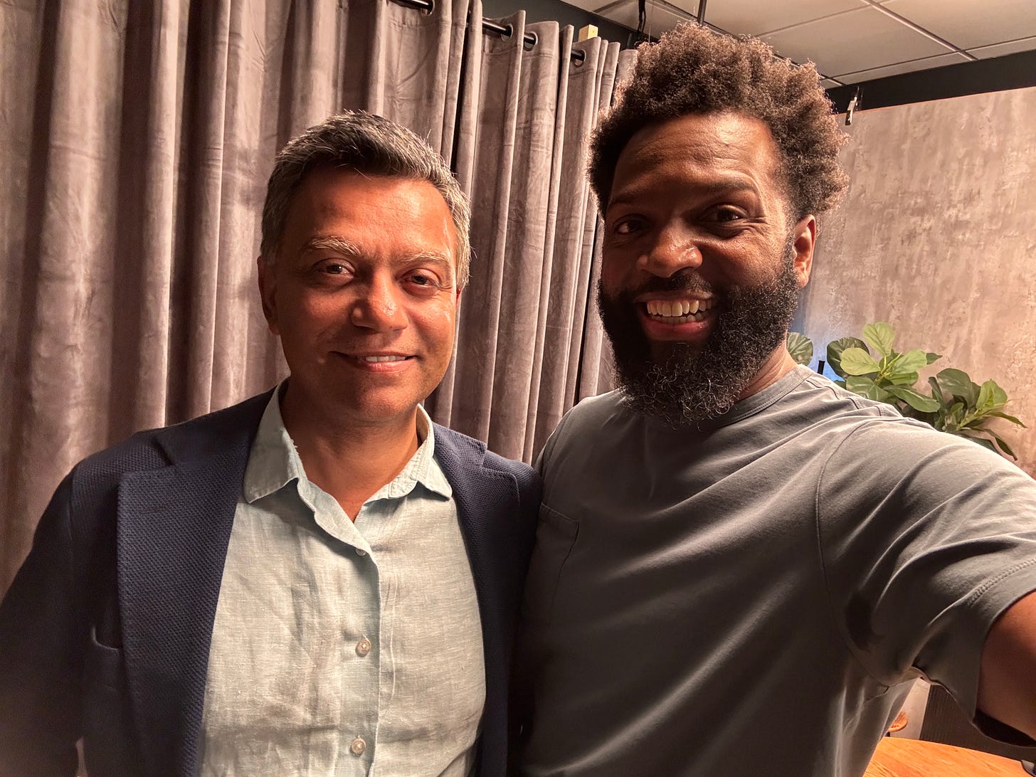 Baratunde Thurston (right), wearing a gray t-shirt, smiles broadly while taking a selfie with Deb Roy (left) dressed in a light blue shirt and dark blazer. They stand in a softly lit studio setting with gray curtains behind them, a textured wall to the side, and a leafy green plant in the background. The atmosphere feels warm, casual, and professional.