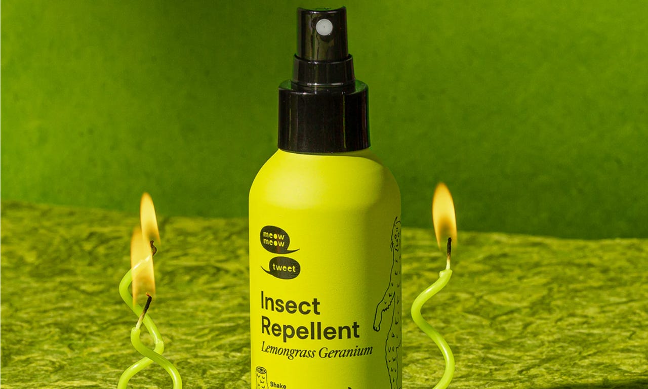 a spray bottle of meow meow tweet's lemongrass geranium insect repellent. the background is different shades and textures of green and lit twisted green candles bracket the bottle.