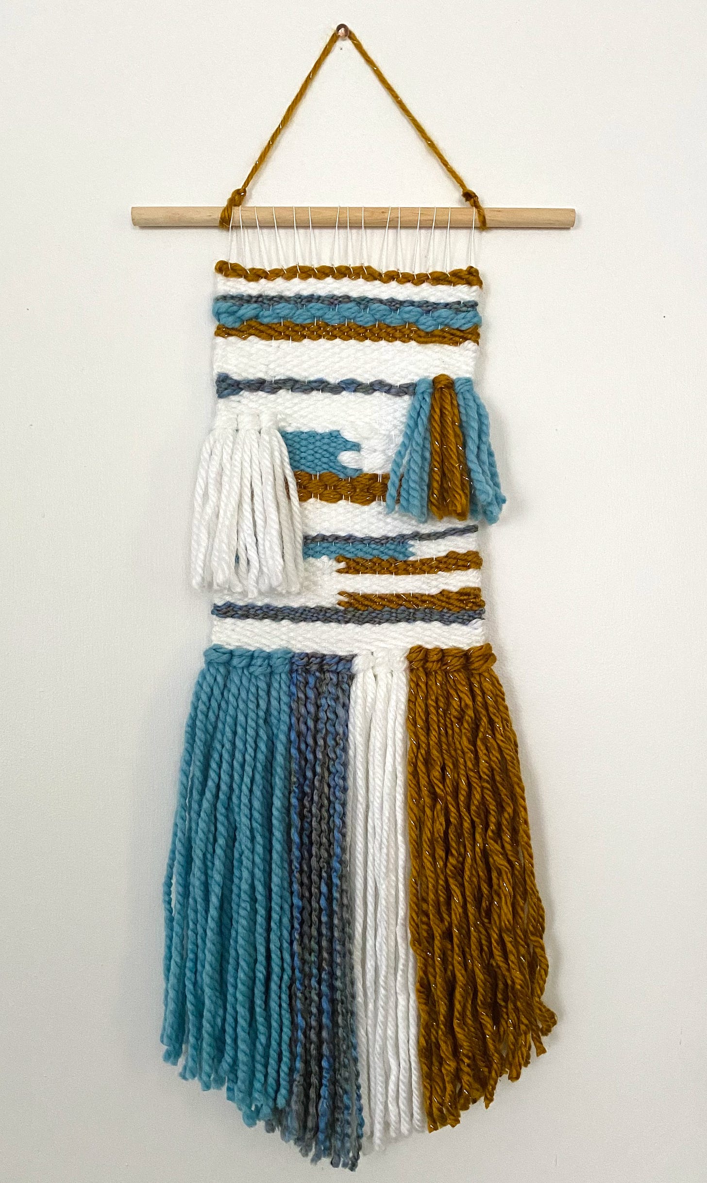 Small wall tapestry weaving with sky blue, deep wavy blue, white, and wavy sparkly rust colored yarn. Hanging on a wooden dowel. The pattern contains fringe and color blocks.