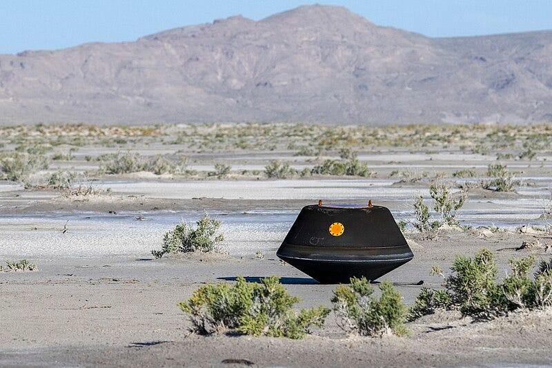 A black space capsule sits perched on the sandy desert floor, surrounded by scrubby brush. Bare, distant mountains fill the horizon in the background.