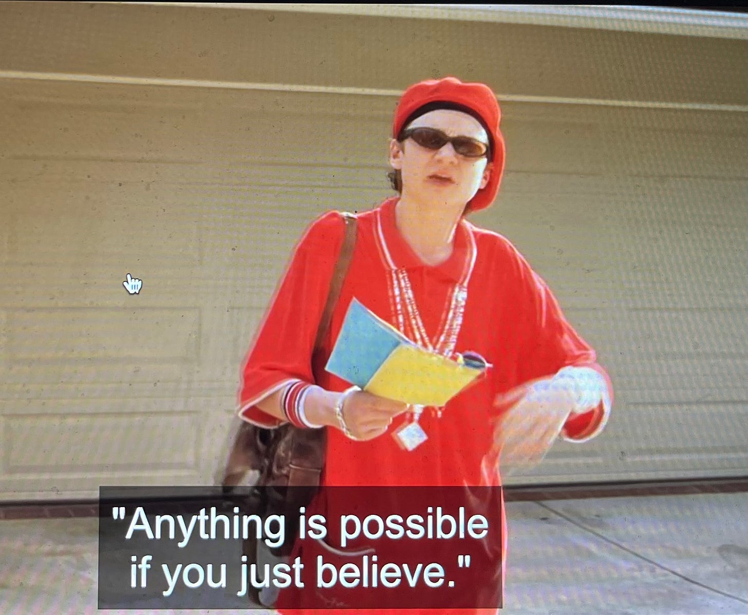 Still from A Cinderella Story showing Carter in his "Snoop Dizzle" look of a red track suit with chains, walking out of his house holding some papers and saying "Anything is possible if you just believe."