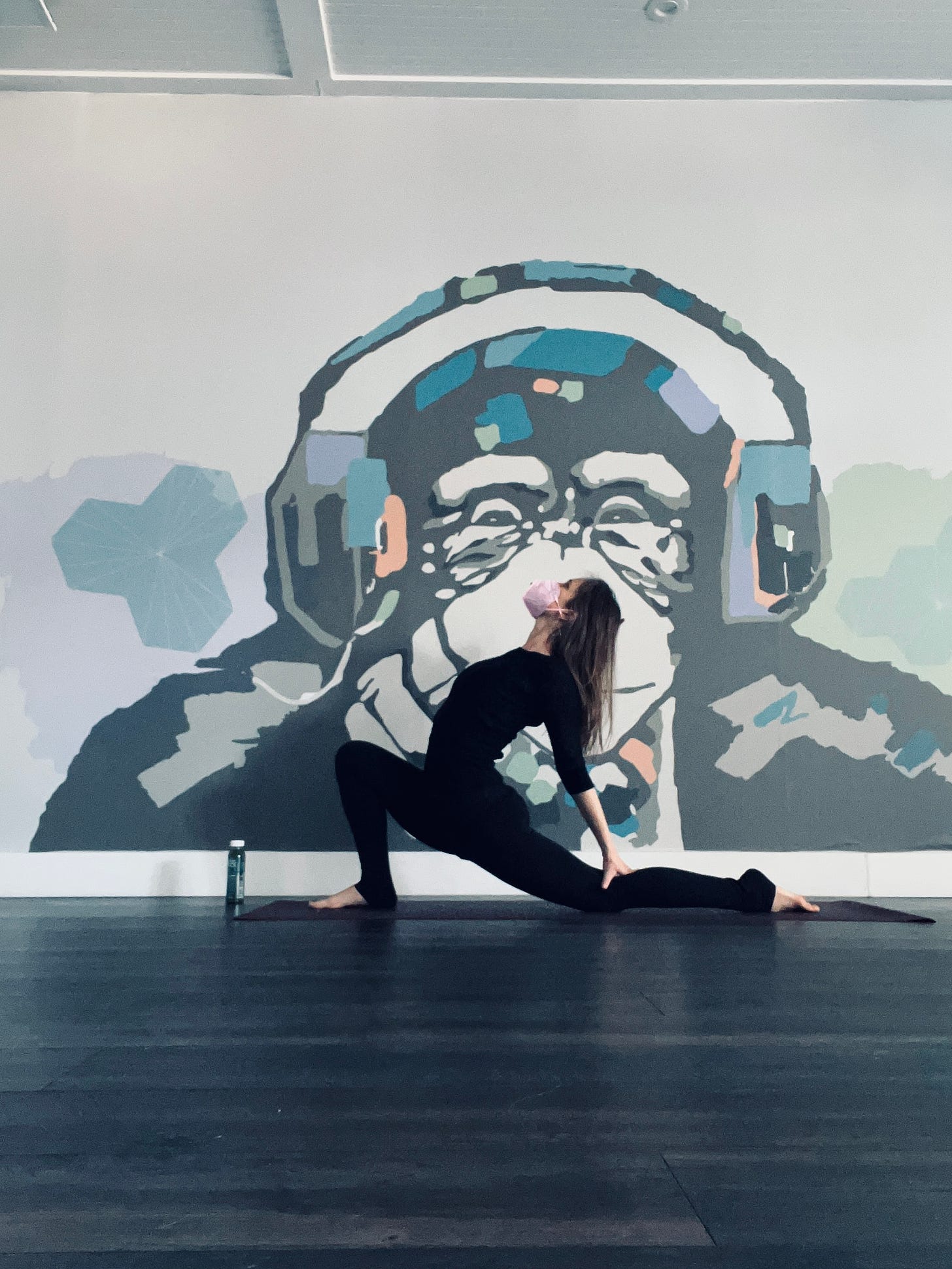 image of the author in a yoga pose wearing all black and a pink mask. behind her on the wall is a painting of a chimpanzee wearing headphones