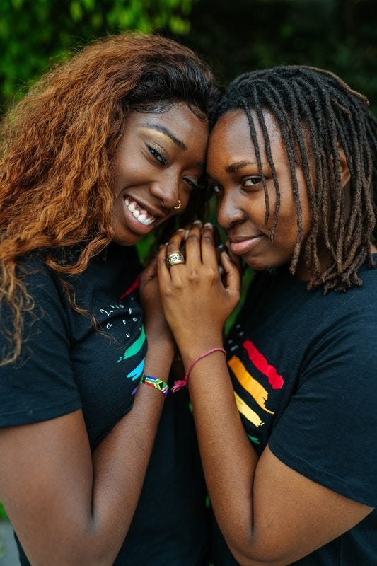 Two Black people standing very close to each other, wearing rainbows, holding hands, and smiling at the camera