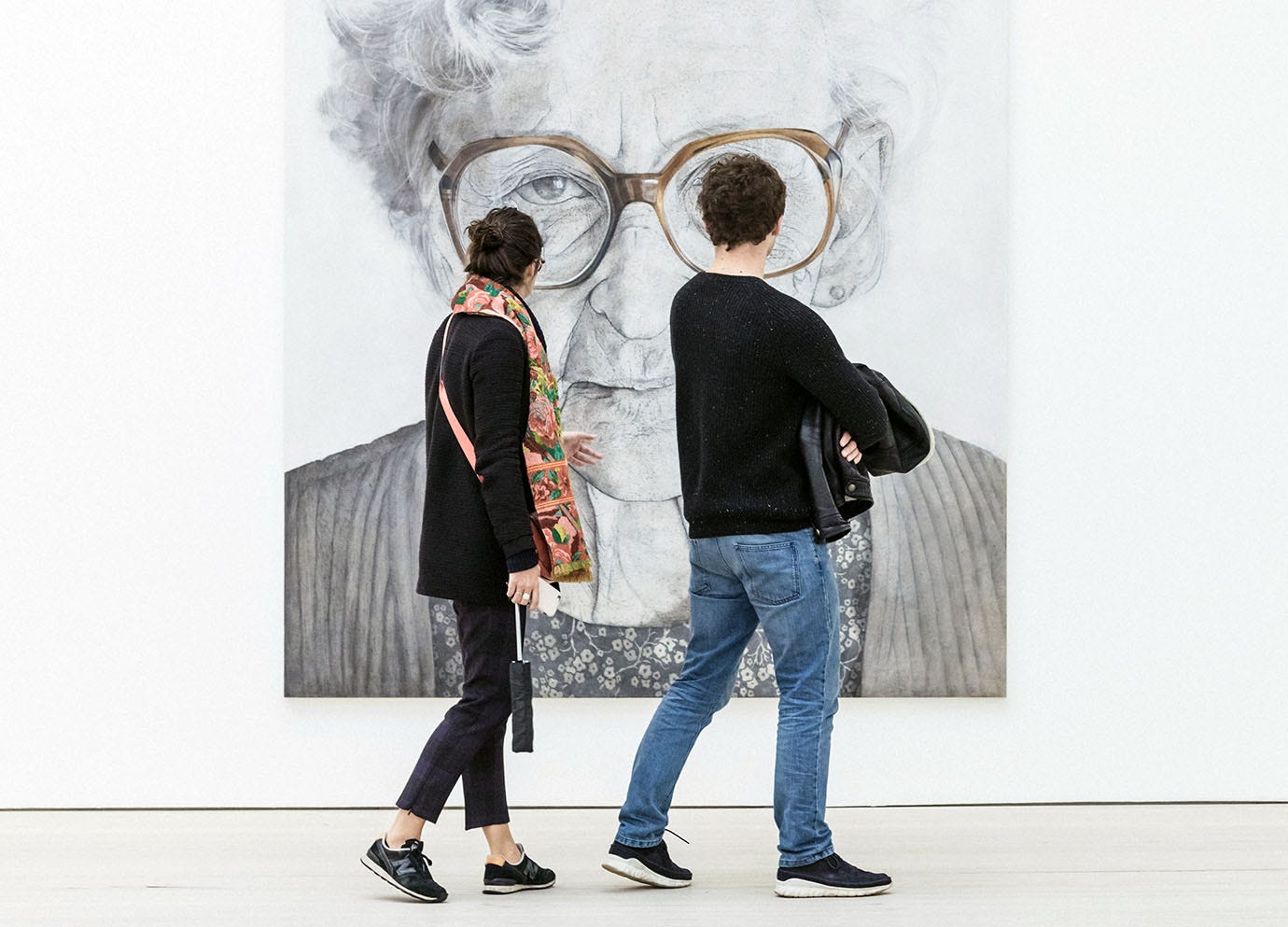 Two people in an art gallery look at a large portrait of an old woman