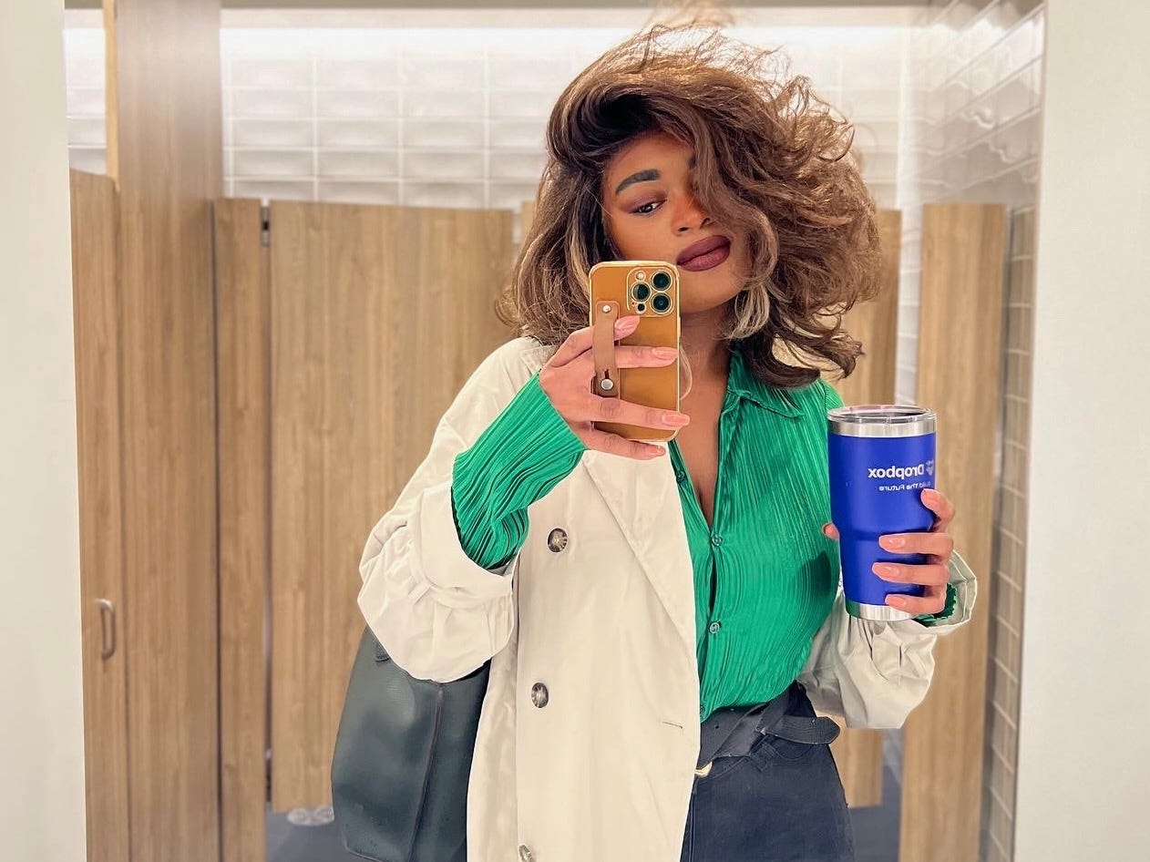 A black woman with short brown hair with brown highlights. She is wearing an emerald green buttoned collar short, a beige Mac jacket & black high waisted jeans while also holding an electric blue Dropbox yeti. 