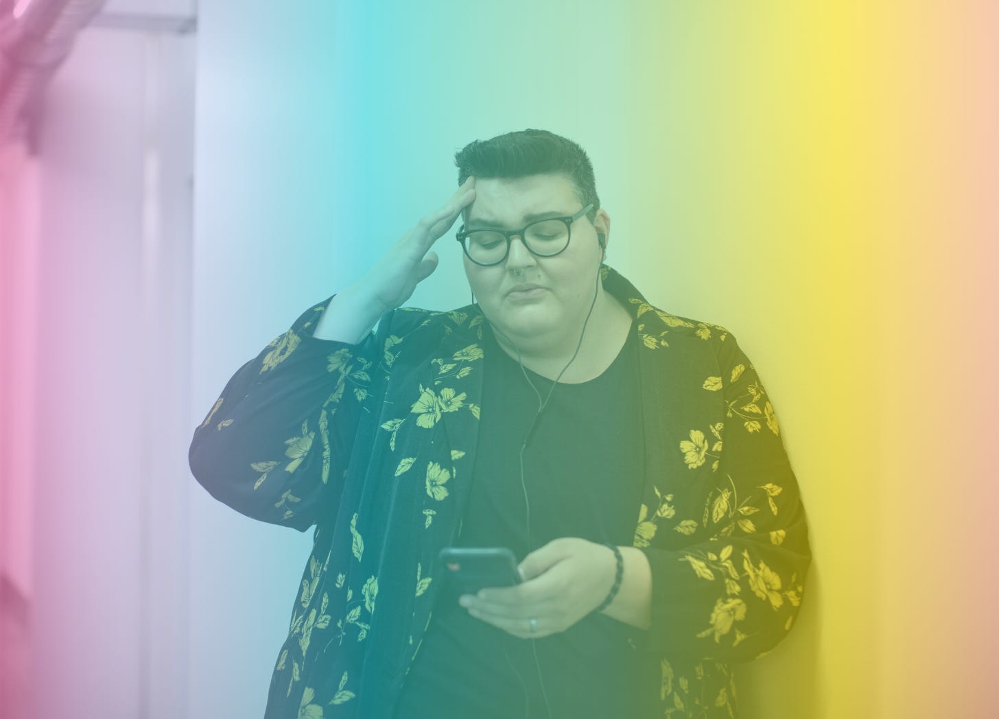 A rainbow-filtered image of a fat, nonbinary individual holding a phone in one hand while grimacing and putting their other hand to their temple