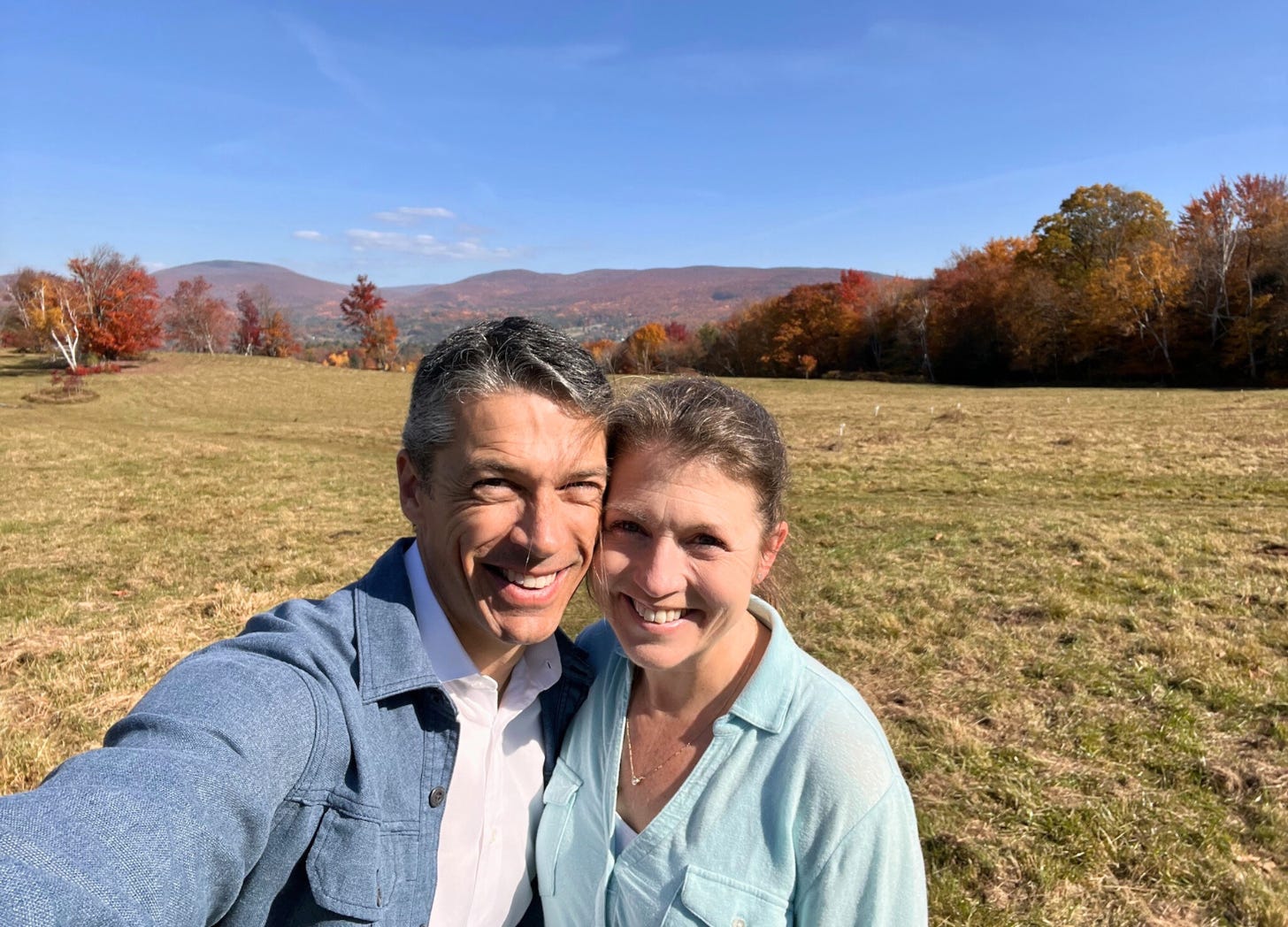 Peter and Amy Julie are smiling for a selfie in an open field on a sunny day, surrounded by colorful autumn trees with mountains in the background.