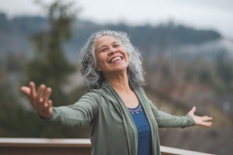 Lively, smiling older woman with long gray hair spreads her arms and hands and looks toward the sky.