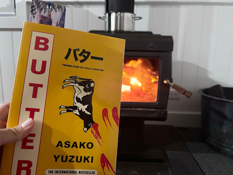 A hand holds a copy of 'Butter' by Asako Yuzuki, in front of a woodfire stove.