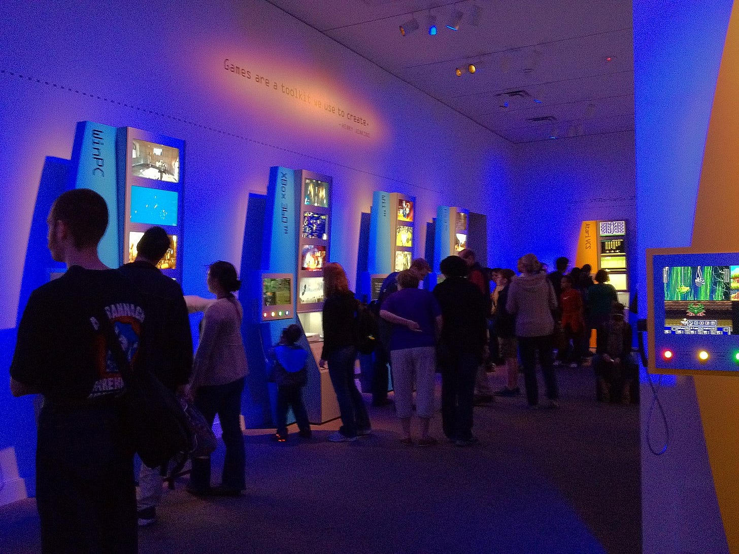 The opening weekend of “The Art of Video Games" exhibit at the Smithsonian American Art Museum in March 2012. Mainly blue and some orange lights bathe visitors looking at screens. A quote above exhibits reads “Games are a toolkit we use to create,” but the attribution is hard to decipher.