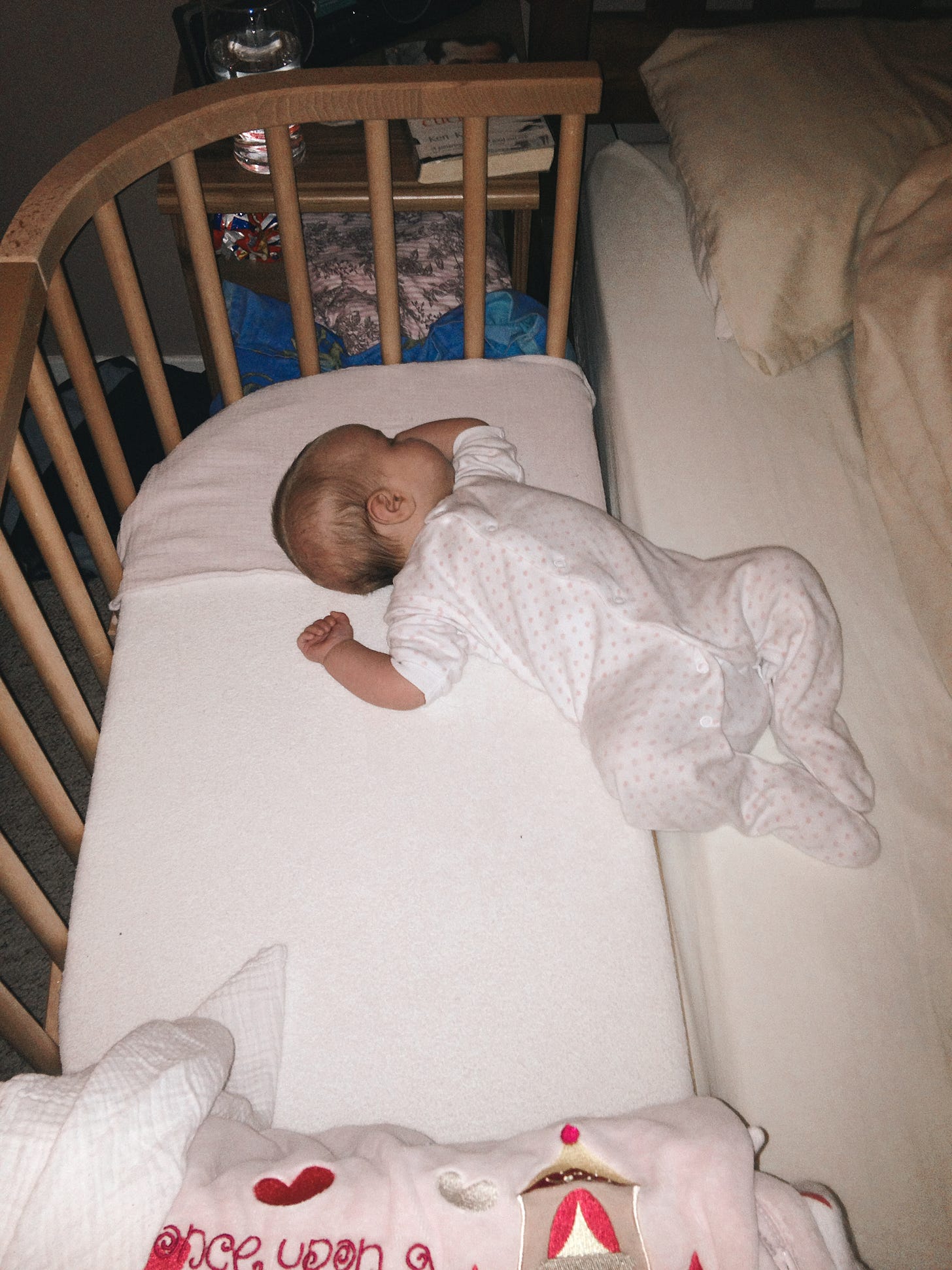 A baby is sleeping mostly on a bedside cot mattress, her feet on the bed