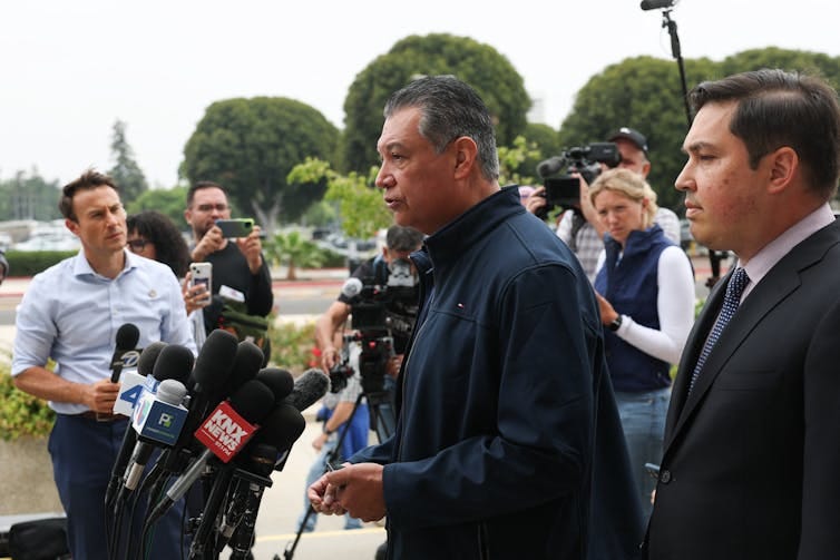 A man speaks in front of a group of microphones, surrounded by other people. A man speaks in front of a group of microphones, surrounded by other people.