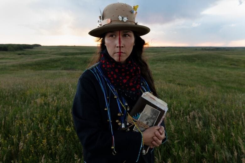 Medium closeup of a woman, the artist Meryl McMaster, standing in a green prairie field under a blue sky, perhaps as twilight approaches. She is facing the viewer, though her gaze looks slightly to the left. A red line is painted down the middle of her face, and she wears a brimmed hat decorted with moths and butterflies and vials of small animal skeletons. In her clasped hands she holds an old diary with a family portrait wrapped to its cover.