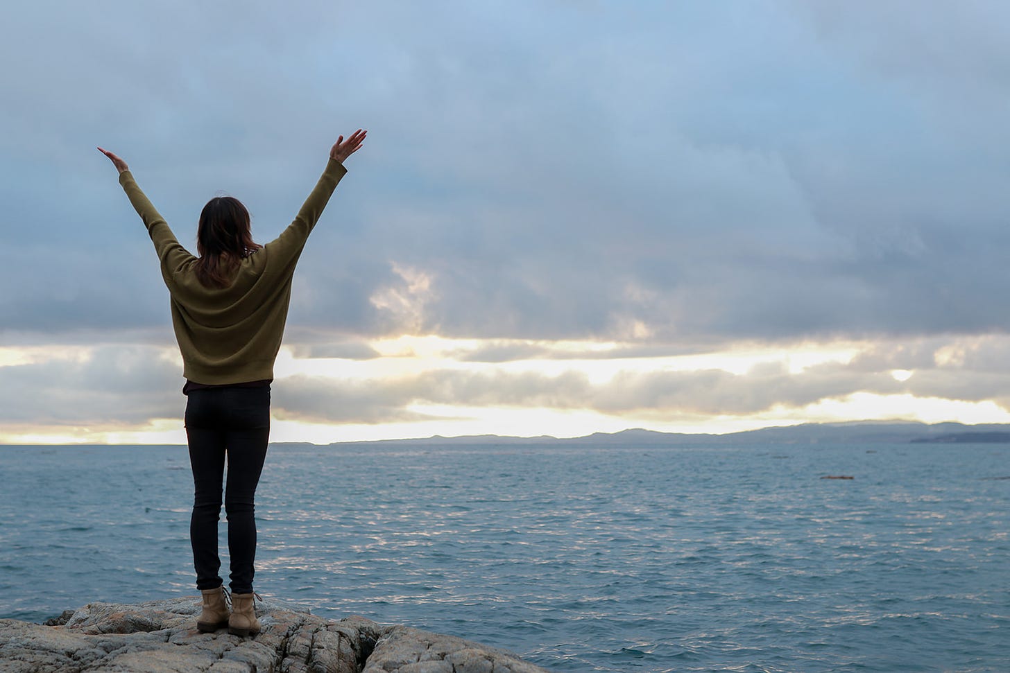 Rayann standing on a rock facing ocean and cloudy sky arms up and outstretched.