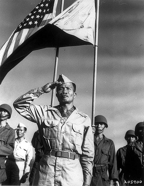 Jose Calugas stands at attention, wearing his Medal. An American flag waves behind him.